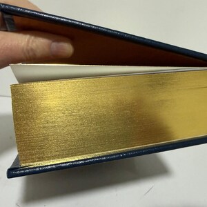 Puede incluir: Un libro de tapa dura con bordes dorados y una cubierta azul marino. Las p&aacute;ginas son de color crema y se ve una cinta dorada como marcap&aacute;ginas. El libro est&aacute; abierto, revelando las p&aacute;ginas interiores y el lado dorado.