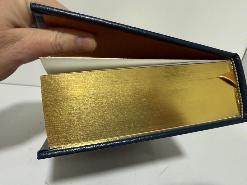 Puede incluir: Un libro de tapa dura con bordes dorados y una cubierta azul marino. Las p&aacute;ginas son de color crema y se ve una cinta dorada como marcap&aacute;ginas. El libro est&aacute; abierto, revelando las p&aacute;ginas interiores y el lado dorado.