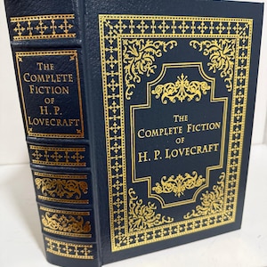 Puede incluir: Un libro de tapa dura titulado "The Complete Fiction of H.P. Lovecraft". El libro tiene una cubierta azul oscuro con detalles y letras doradas. El lomo y la cubierta presentan dise&ntilde;os y textos dorados ornamentados.