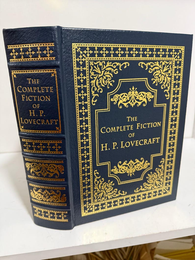 Puede incluir: Un libro de tapa dura titulado "The Complete Fiction of H.P. Lovecraft". El libro tiene una cubierta azul oscuro con detalles y letras doradas. El lomo y la cubierta presentan dise&ntilde;os y textos dorados ornamentados.