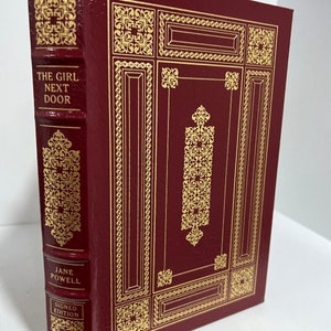 May include: A burgundy leather-bound book with gold accents and the title "The Girl Next Door" by Jane Powell. The book is a signed edition.