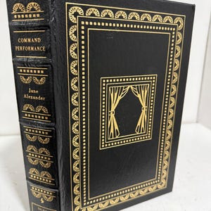 Puede incluir: Libro de tapa dura negro, "Command Performance" de Jane Alexander. Cuenta con detalles dorados y texto. El lomo muestra el título, autor y "Signed First Edition".