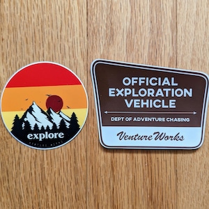 Könnte beinhalten: Zwei runde Aufkleber. Ein Aufkleber zeigt eine Bergkette mit einer untergehenden Sonne und dem Text "explore". Der andere Aufkleber ist braun mit weißem Text, der "OFFICIAL EXPLORATION VEHICLE DEPT OF ADVENTURE CHASING Venture Works" lautet.