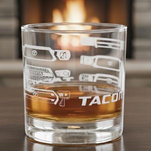 May include: A clear glass tumbler with a white graphic design and the word "TACO" in bold letters. The glass is filled with a golden-brown liquid. The background features a fireplace with a lit fire.
