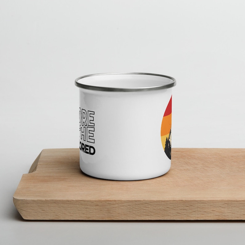 May include: White enamel mug with a silver rim. The mug features the text "Explore the Unexplored" in black, stacked vertically. A circular graphic on the right side depicts a mountain silhouette against a sunset gradient of red, orange, and yellow.