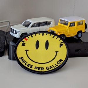 May include: A yellow smiley face patch with the text "Smiles Per Gallon" and a red needle pointing to the "F" on the gauge. The patch is sitting on top of a black surface with two toy cars, one white and one yellow.