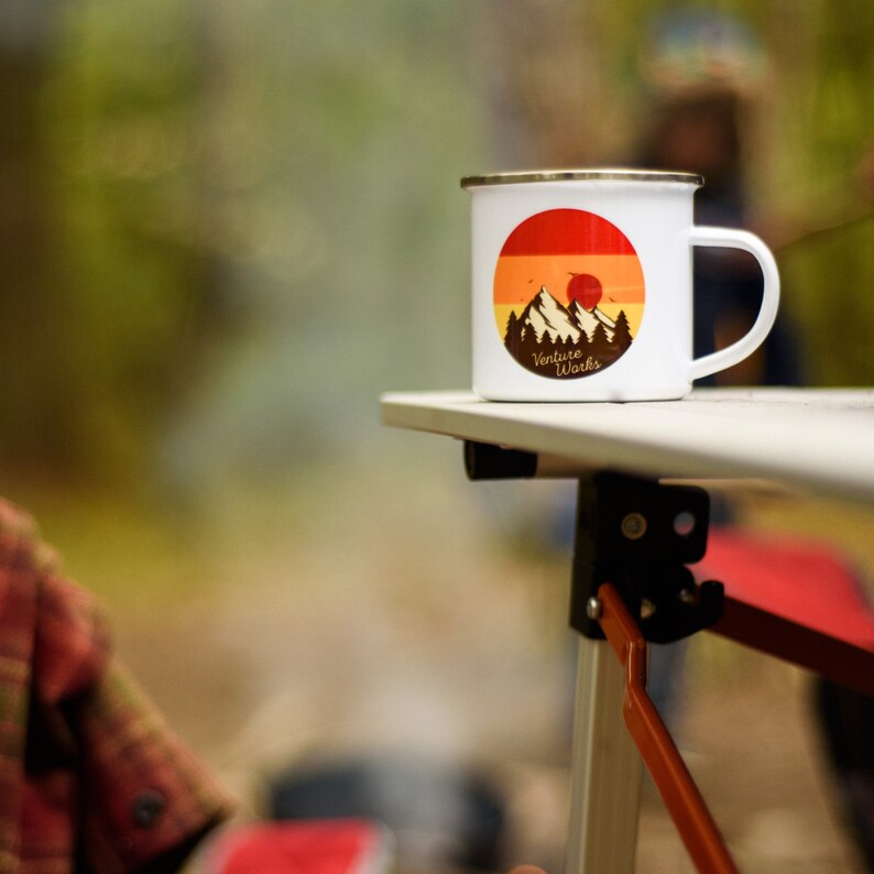 May include: White enamel camping mug with a gold rim, featuring a mountain and sunset graphic with the text "Venture Works." The mug is sitting on a light-colored table, suggesting an outdoor setting.