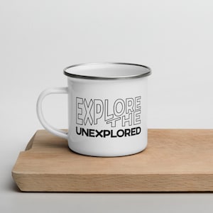 May include: White enamel mug with a black rim and handle. The mug features the text "EXPLORE THE UNEXPLORED" in a bold, outlined font. The mug is sitting on a wooden cutting board.