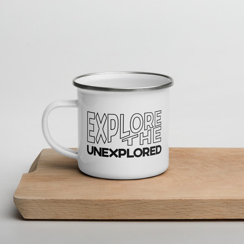 May include: White enamel mug with a black rim and handle. The mug features the text "EXPLORE THE UNEXPLORED" in a bold, outlined font. The mug is sitting on a wooden cutting board.