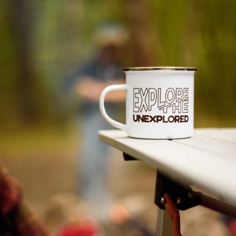 May include: White enamel camping mug with a black rim and handle. The mug features the text "EXPLORE THE UNEXPLORED" in a bold, black font. The mug is sitting on a white table, with a blurred outdoor background.