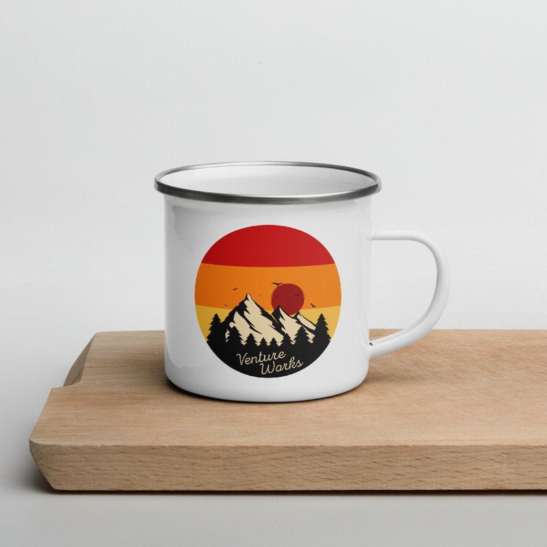 May include: White enamel mug with a black rim and handle. The mug features a circular design with a mountain range, trees, and a sunset in shades of red, orange, and yellow. The text "Venture Works" is below the mountains.