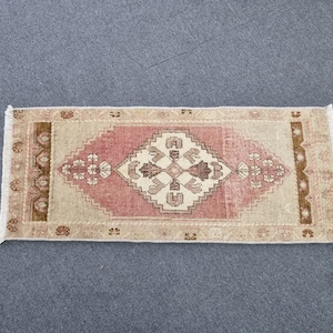 May include: A small, rectangular rug with a pink and beige geometric pattern. The rug has a fringe border and is made of wool.