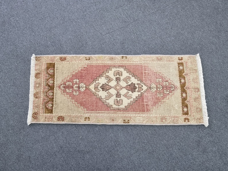 May include: A small, rectangular rug with a pink and beige geometric pattern. The rug has a fringe border and is made of wool.