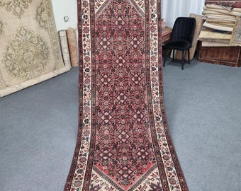 May include: A long, narrow rug with a cream background and a red, black, and blue geometric pattern. The rug has a floral border and is fringed on the edges.