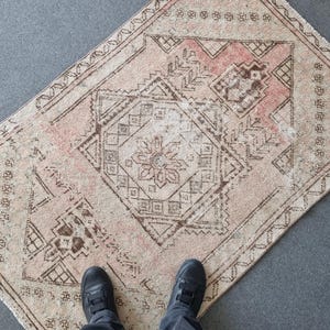 May include: A rectangular, vintage-style rug with a faded, distressed appearance. The rug features a central diamond pattern with floral and geometric motifs in shades of beige, brown, and faded rose. The rug is placed on a gray surface.