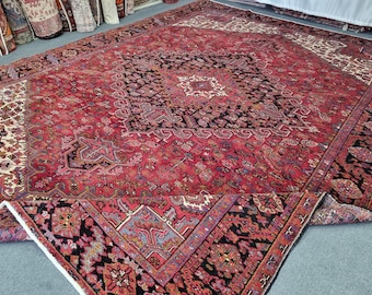 May include: A large, rectangular, red Persian rug with an intricate geometric pattern. The rug features a central medallion design in black and cream, surrounded by detailed borders and floral motifs. The rug is partially rolled up, revealing its underside.