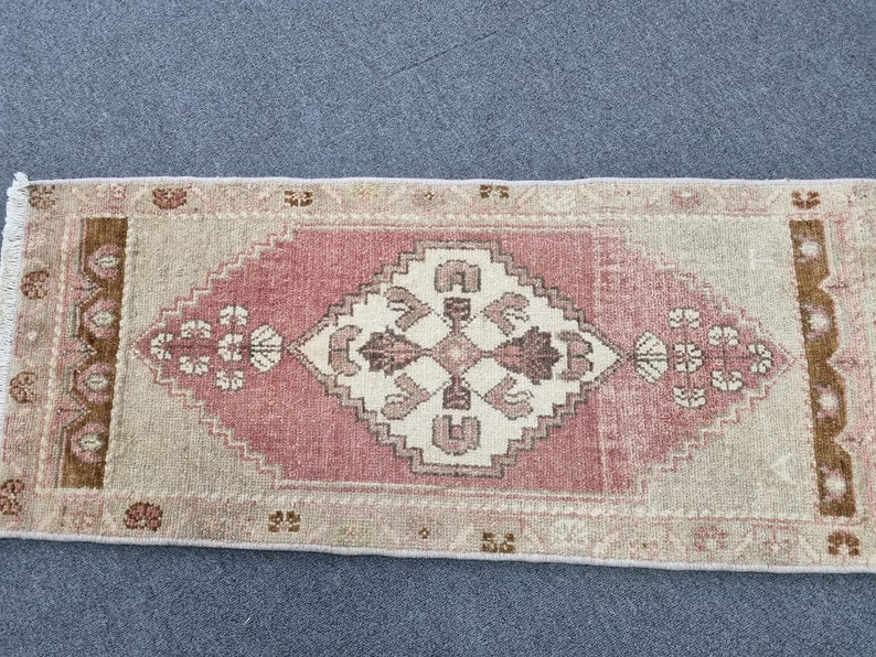 May include: A pink and beige runner rug with a white geometric pattern in the center. The rug has a fringe border.