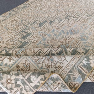 May include: A rolled-up, beige area rug with an intricate geometric pattern in shades of blue, green, and brown. The rug's design features repeating shapes and floral motifs, creating a vintage or antique aesthetic. The rug is made of a woven material.