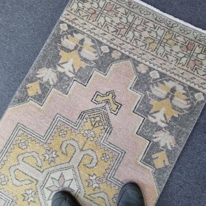 Puede incluir: Una alfombra de color rosa y gris con un dise&ntilde;o geom&eacute;trico amarillo y blanco en el centro. La alfombra tiene un borde de flecos.
