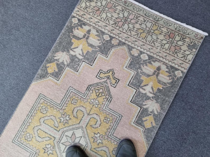 Puede incluir: Una alfombra de color rosa y gris con un dise&ntilde;o geom&eacute;trico amarillo y blanco en el centro. La alfombra tiene un borde de flecos.