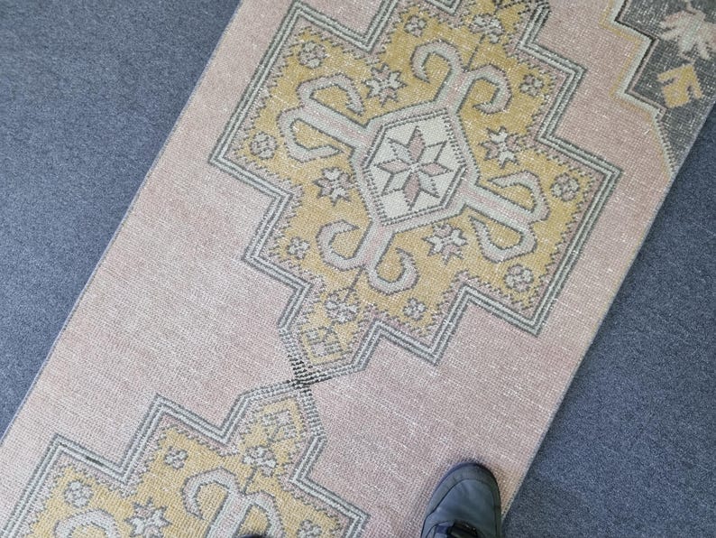Puede incluir: Una alfombra de color rosa claro y amarillo con un dise&ntilde;o geom&eacute;trico. La alfombra tiene un aspecto descolorido y est&aacute; hecha de un material suave y tejido.