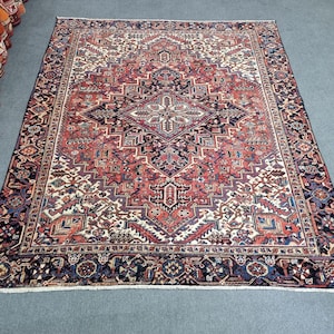 May include: A large, rectangular rug with an intricate, geometric pattern. The rug features a central diamond design in shades of red, navy, and cream, surrounded by a detailed border. The rug's dimensions are not visible in the image.