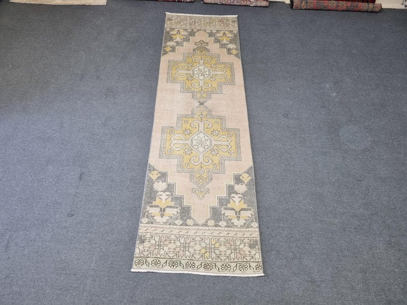 Puede incluir: Una alfombra rectangular larga con fondo crema y un patr&oacute;n geom&eacute;trico en tonos amarillos y grises. El patr&oacute;n presenta una forma de diamante central con cuatro diamantes m&aacute;s peque&ntilde;os dentro.