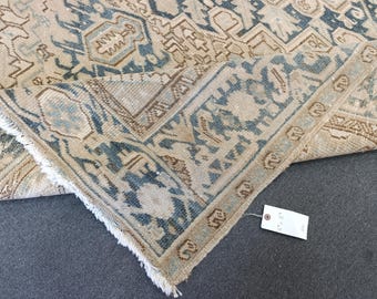 May include: A section of a beige and blue patterned rug with a distressed, vintage look. The rug features geometric and floral designs in shades of blue, brown, and beige. The rug's edge is visible, showing a fringed border. A tag is attached to the rug.