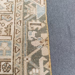 May include: Close-up of a section of a rug with a detailed, geometric pattern. The rug features a combination of beige, light blue, and olive green hues. The design includes repeating shapes and borders, with a textured appearance.