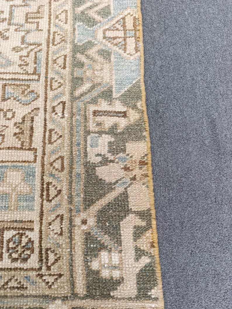 May include: Close-up of a section of a rug with a detailed, geometric pattern. The rug features a combination of beige, light blue, and olive green hues. The design includes repeating shapes and borders, with a textured appearance.