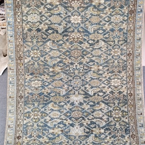 May include: A rectangular area rug featuring a complex, repeating geometric and floral pattern in shades of dusty blue, beige, cream, and brown. The rug has a detailed border with linear motifs and a dense, symmetrical design throughout the center.
