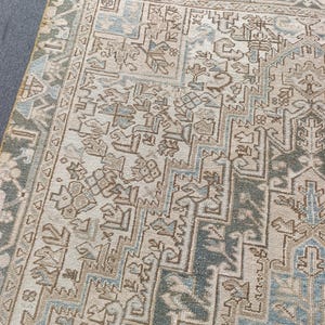May include: An area rug with an intricate, geometric pattern in shades of beige, blue, and green. The rug has a distressed, vintage look, with a detailed border and central design. The rug is rectangular and appears to be made of woven material.