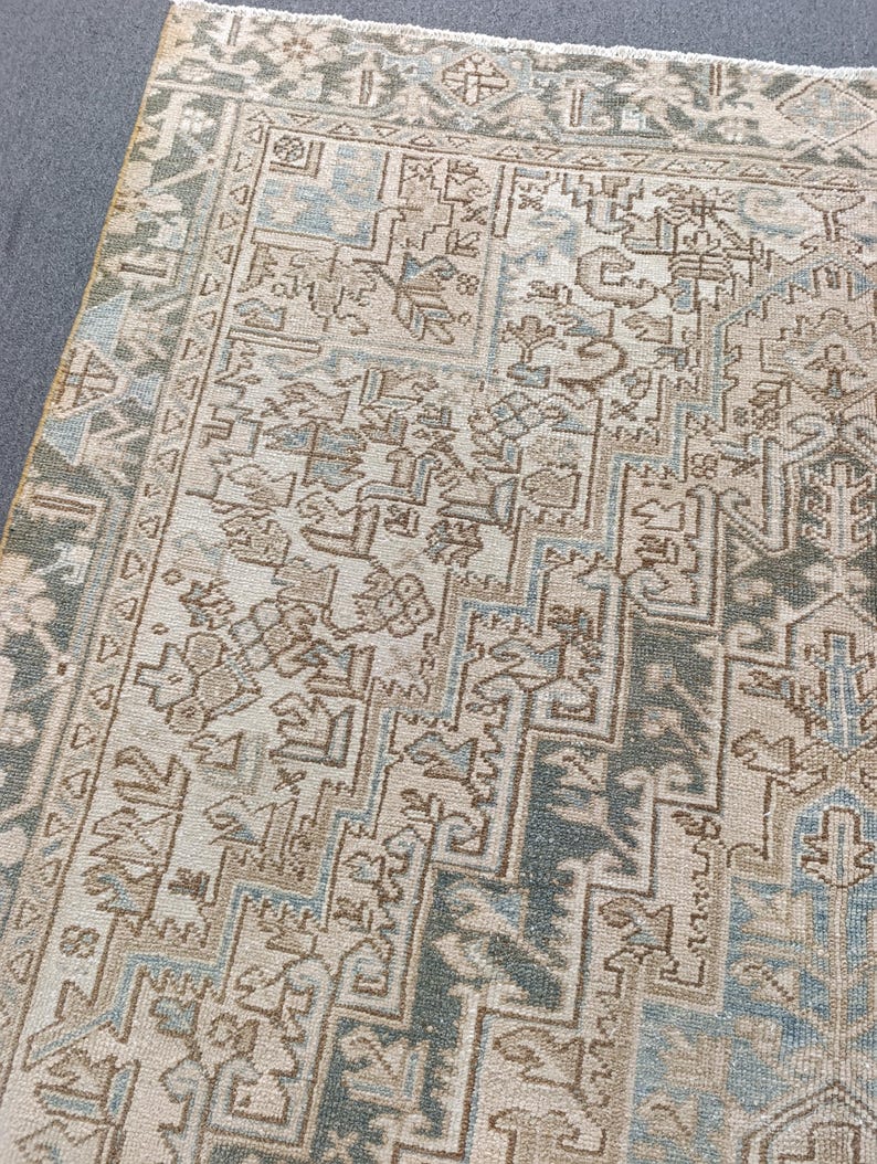 May include: An area rug with an intricate, geometric pattern in shades of beige, blue, and green. The rug has a distressed, vintage look, with a detailed border and central design. The rug is rectangular and appears to be made of woven material.
