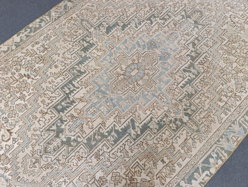 May include: A rectangular area rug with an intricate, geometric pattern. The rug features a central medallion in shades of blue and beige, surrounded by a border of teal and beige. The overall design is symmetrical and detailed, with a vintage aesthetic.