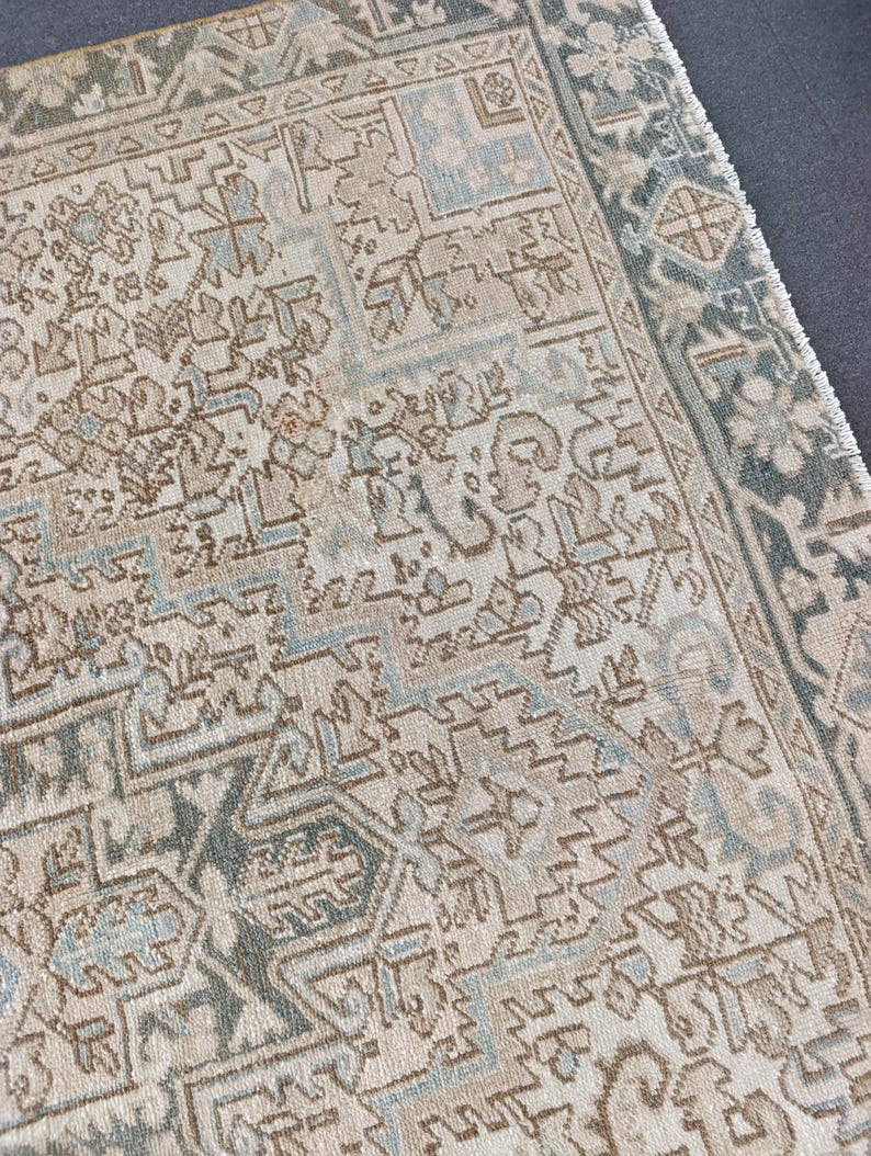 May include: Close-up of a section of a vintage rug with an intricate geometric pattern. The rug features a cream-colored background with detailed designs in shades of brown, blue, and green. The edges have a darker green border with a fringe.