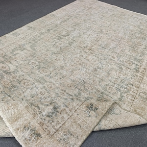 May include: A rectangular area rug featuring a faded, distressed pattern in shades of beige, cream, and muted sage green. The design includes intricate floral and geometric motifs with a wide border, and one corner is folded over to show the underside.