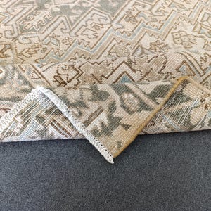 May include: A beige and brown patterned rug with a folded corner, revealing the underside. The rug features a geometric design with accents of blue and green. The edges are finished with a white fringe.
