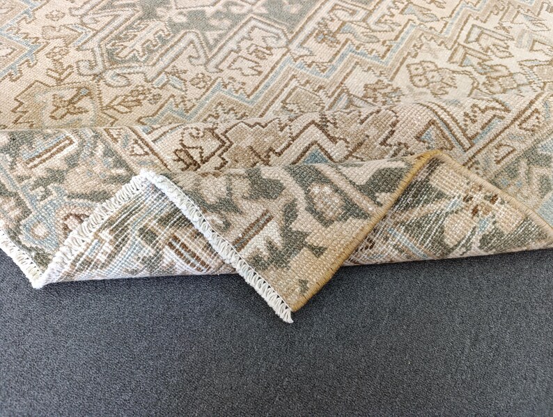 May include: A beige and brown patterned rug with a folded corner, revealing the underside. The rug features a geometric design with accents of blue and green. The edges are finished with a white fringe.
