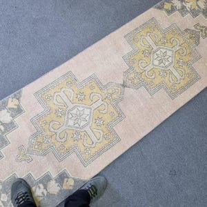 Puede incluir: Una alfombra de pasillo rosa y crema con un patr&oacute;n geom&eacute;trico en amarillo y gris. La alfombra tiene un aspecto desgastado y est&aacute; hecha de un material suave y tejido.