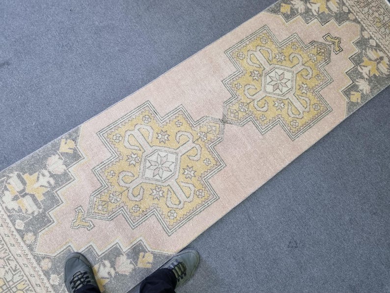 Puede incluir: Una alfombra de pasillo rosa y crema con un patr&oacute;n geom&eacute;trico en amarillo y gris. La alfombra tiene un aspecto desgastado y est&aacute; hecha de un material suave y tejido.