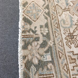 May include: Close-up of a beige and gray patterned rug with a fringe edge. The rug features geometric designs in shades of gray, blue, and brown. The rug is placed next to a gray surface.