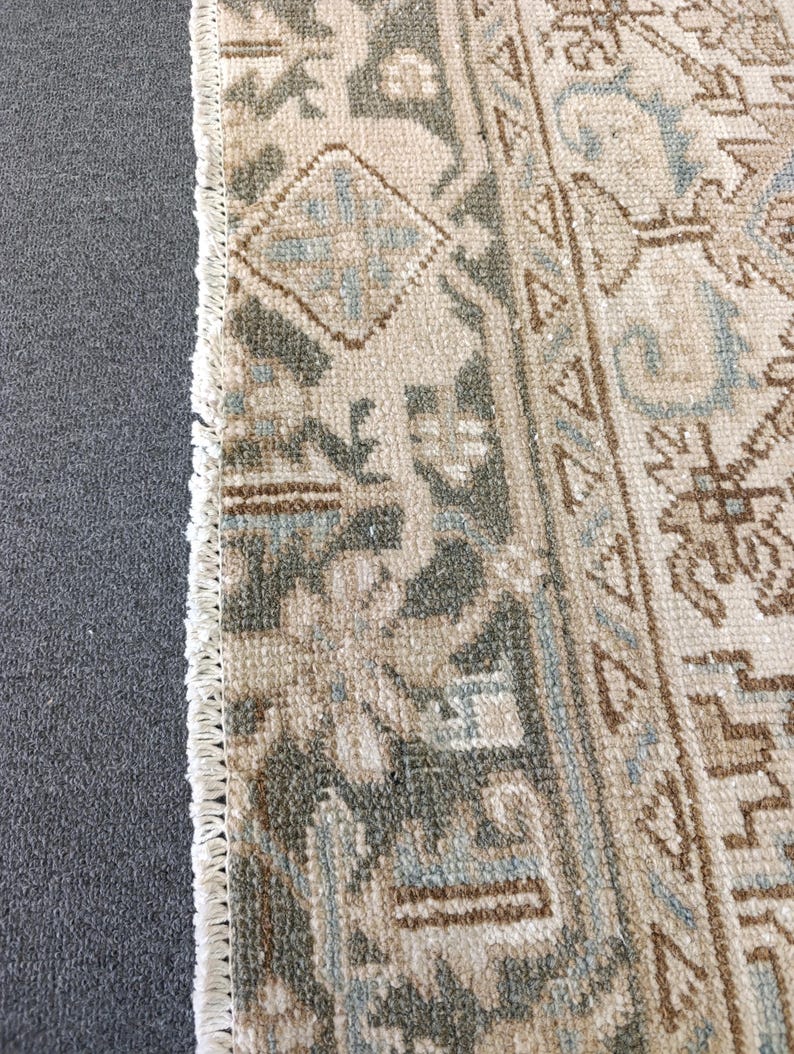 May include: Close-up of a beige and gray patterned rug with a fringe edge. The rug features geometric designs in shades of gray, blue, and brown. The rug is placed next to a gray surface.