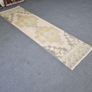 Puede incluir: Una alfombra de pasillo vintage con fondo crema y un patr&oacute;n geom&eacute;trico en tonos amarillos y grises. La alfombra tiene un borde con flecos.