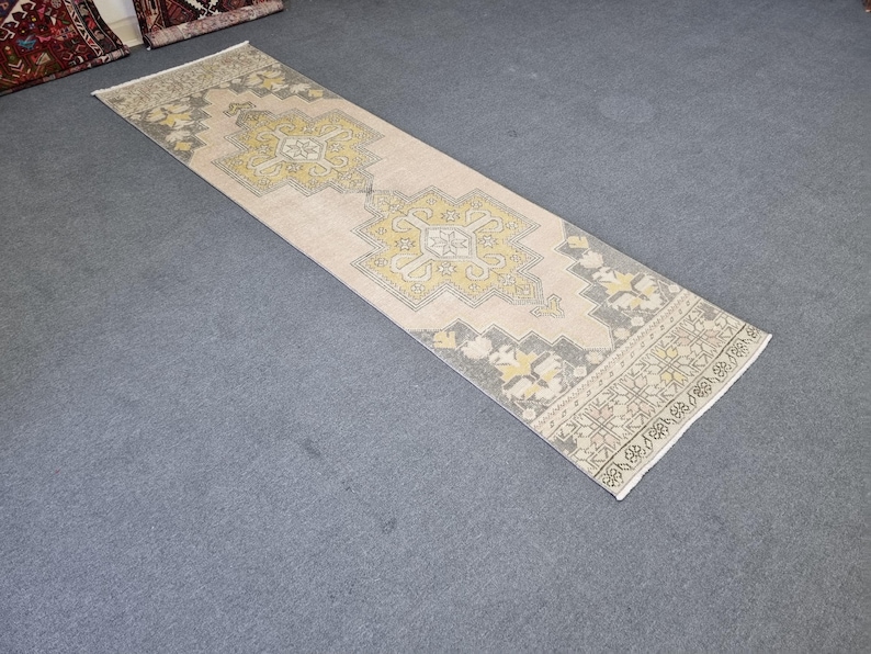 Puede incluir: Una alfombra de pasillo vintage con fondo crema y un patr&oacute;n geom&eacute;trico en tonos amarillos y grises. La alfombra tiene un borde con flecos.