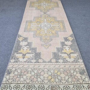 Puede incluir: Una alfombra larga y estrecha con un patr&oacute;n geom&eacute;trico rosa, gris y amarillo. La alfombra tiene un borde de flecos.
