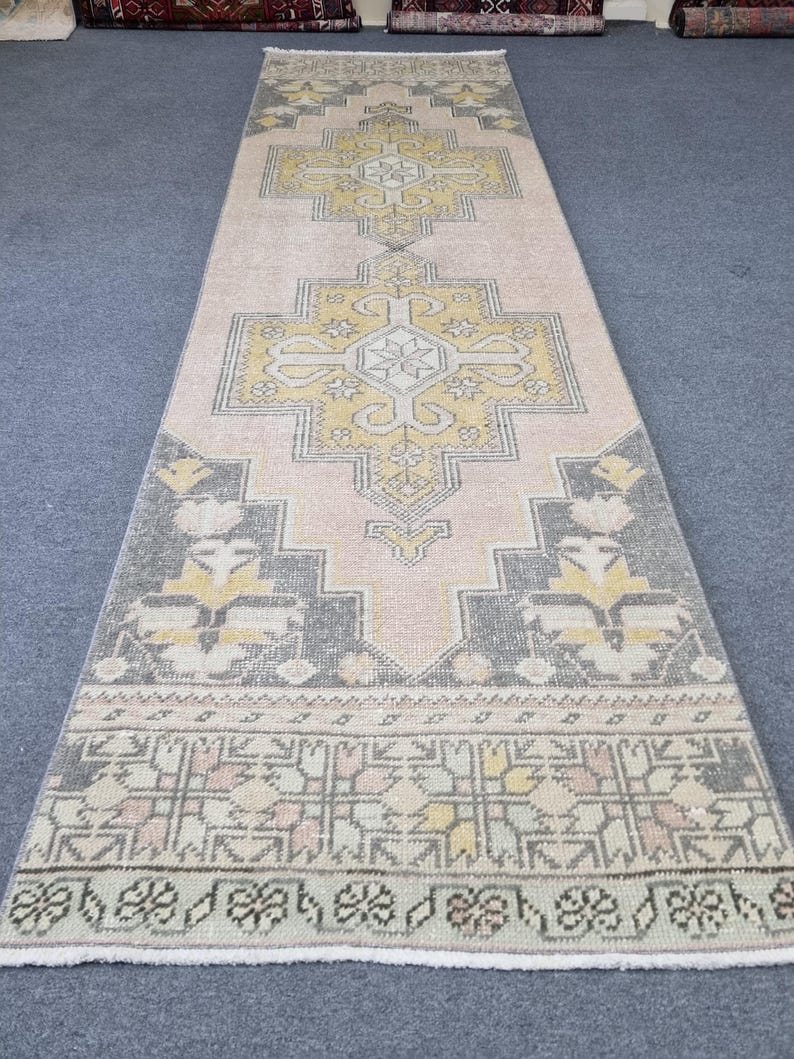 Puede incluir: Una alfombra larga y estrecha con un patr&oacute;n geom&eacute;trico rosa, gris y amarillo. La alfombra tiene un borde de flecos.