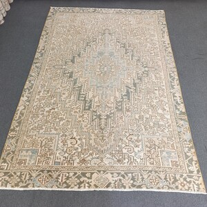 May include: A rectangular, beige rug with an intricate geometric pattern in shades of green, brown, and blue. The rug features a central design with a border, creating a traditional aesthetic. The rug is placed on a gray surface.