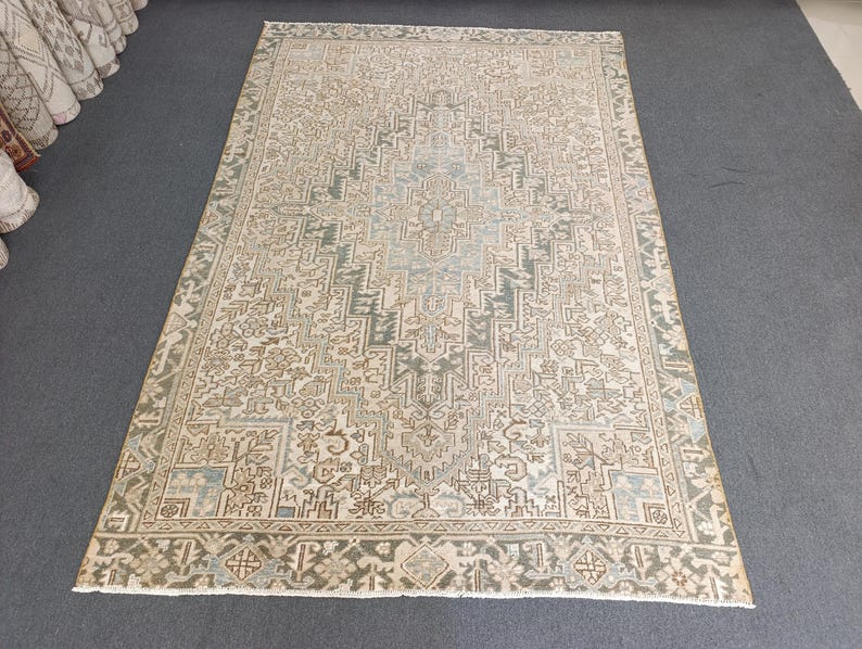May include: A rectangular, beige rug with an intricate geometric pattern in shades of green, brown, and blue. The rug features a central design with a border, creating a traditional aesthetic. The rug is placed on a gray surface.