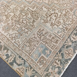 May include: A corner of a vintage rug with a detailed geometric pattern. The rug features a cream-colored background with intricate designs in shades of brown, blue, and green. The border has a darker green hue, and the rug has a fringed edge.