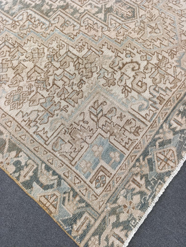 May include: A corner of a vintage rug with a detailed geometric pattern. The rug features a cream-colored background with intricate designs in shades of brown, blue, and green. The border has a darker green hue, and the rug has a fringed edge.